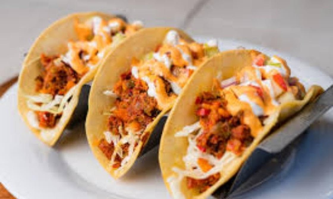 Three tacos filled with seasoned ground meat, shredded lettuce, diced tomatoes, and drizzled with orange sauce and sour cream, on a white plate.