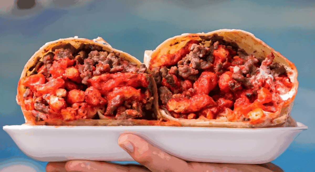 A close-up of a burrito cut in half, filled with spicy red Cheetos, ground beef, and cheese, held on a plate against a blue sky background.
