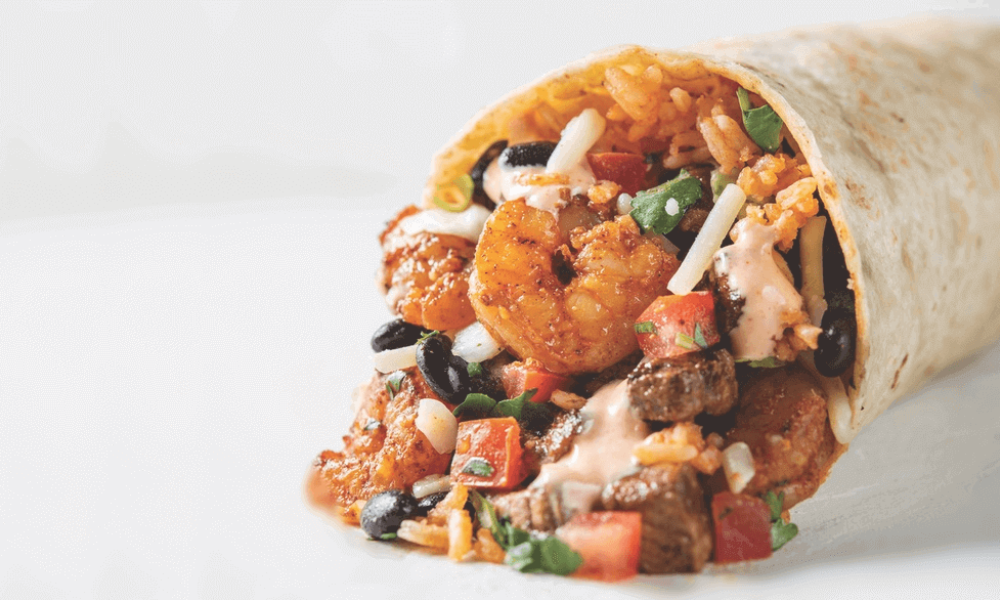 A close-up of a burrito with shrimp, beef, rice, black beans, cheese, tomatoes, and cilantro, wrapped in a tortilla on a white surface.