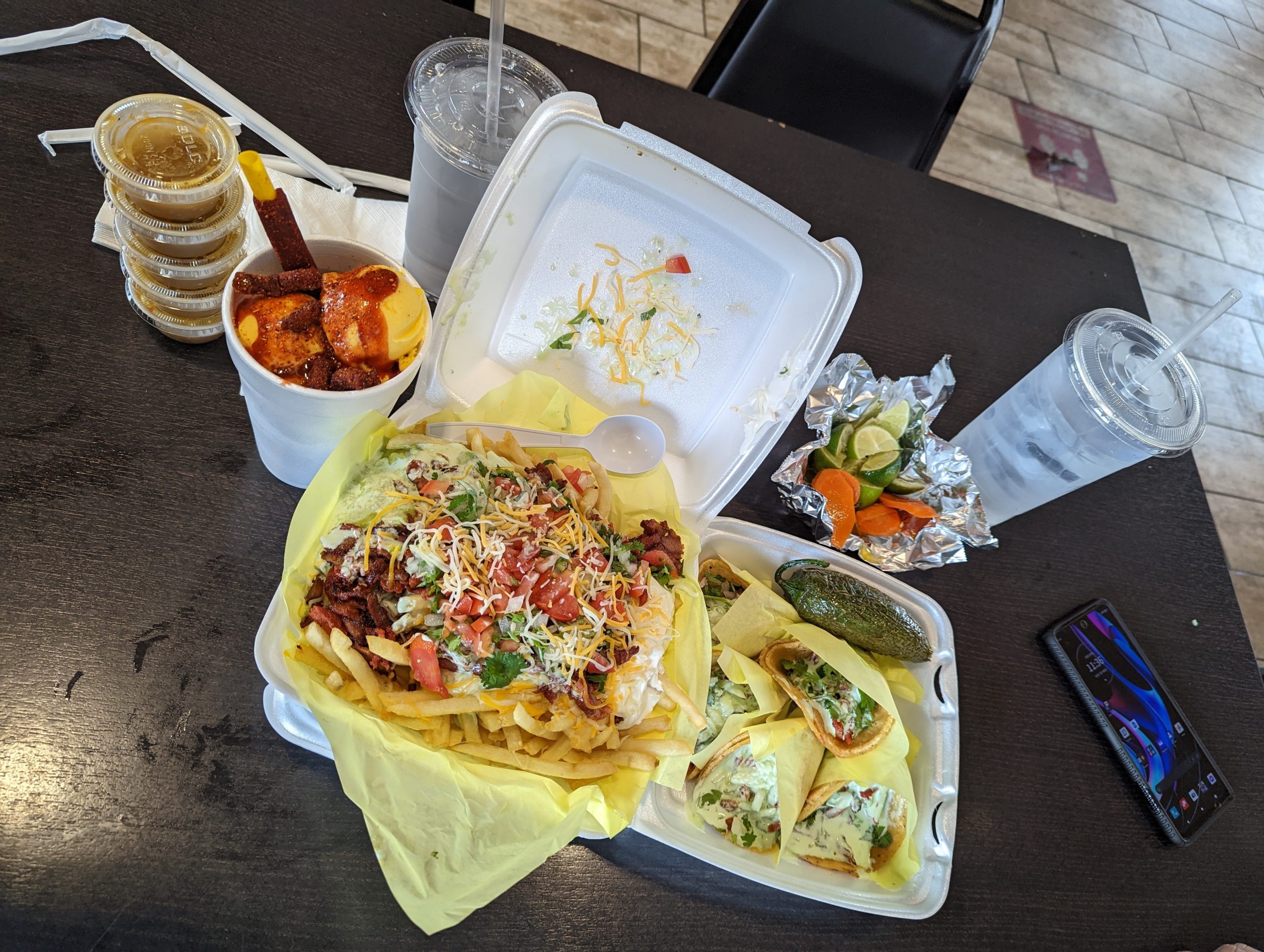 A table with loaded fries topped with meat, cheese, and veggies in a styrofoam tray, tacos, a cup with chamoy sauce, sauces, beverages, and garnish.