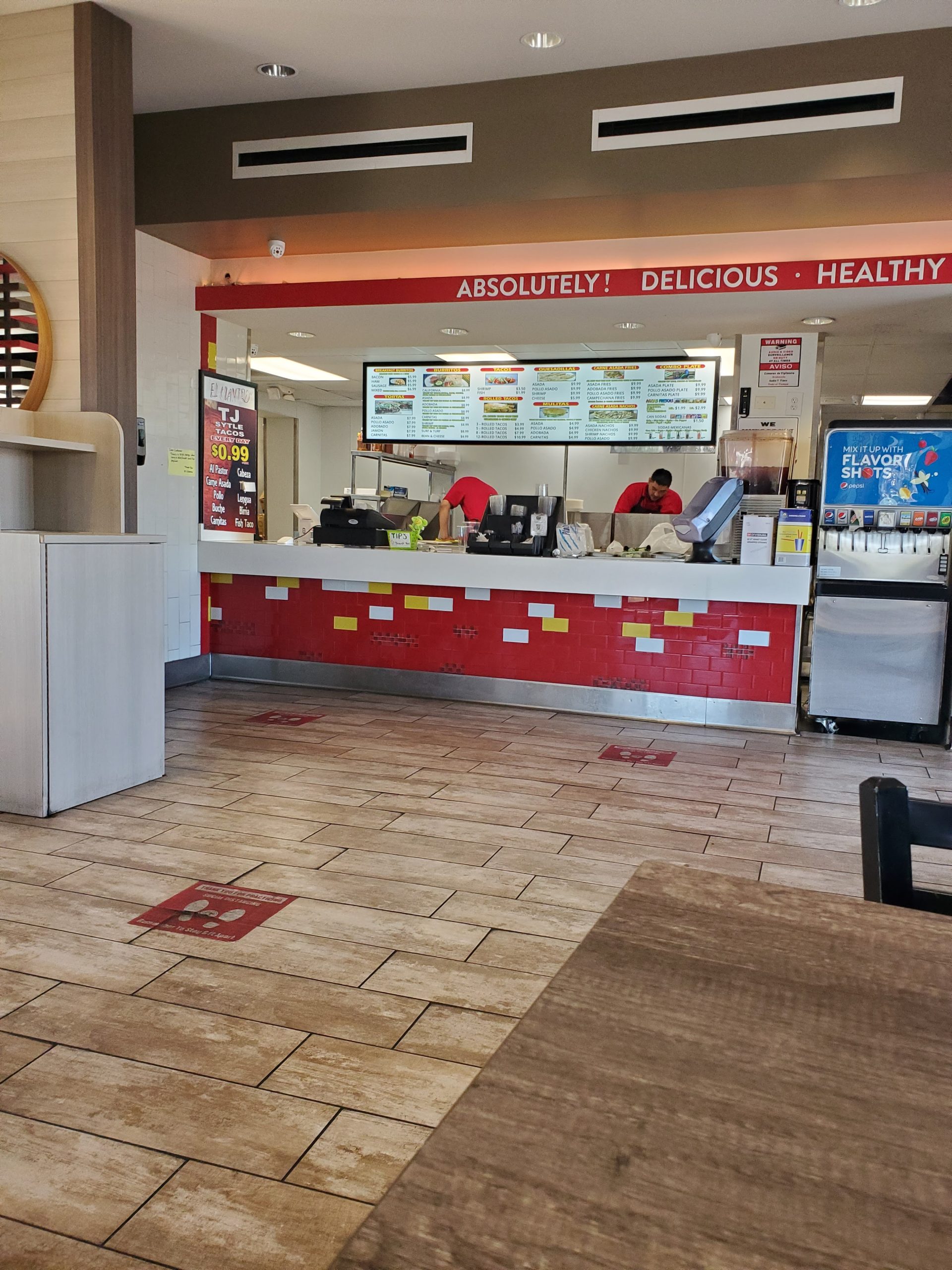 Inside a fast-food restaurant with a red and white counter. Two staff in red shirts work behind it. Menus overhead; wood flooring enhances the casual ambiance.